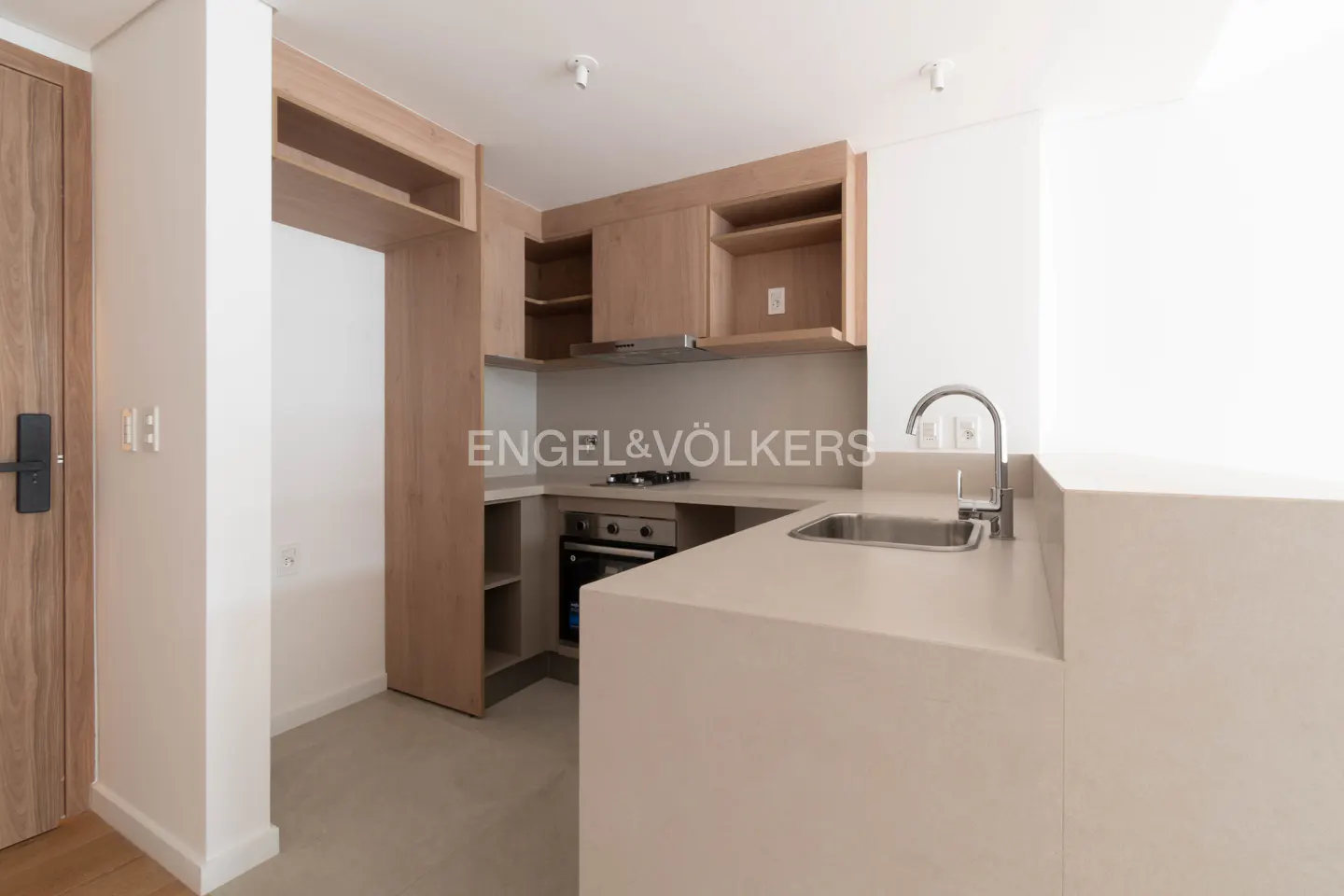 Modern kitchen with light wood cabinets, beige countertops, stainless steel sink, and built-in oven in a bright room.