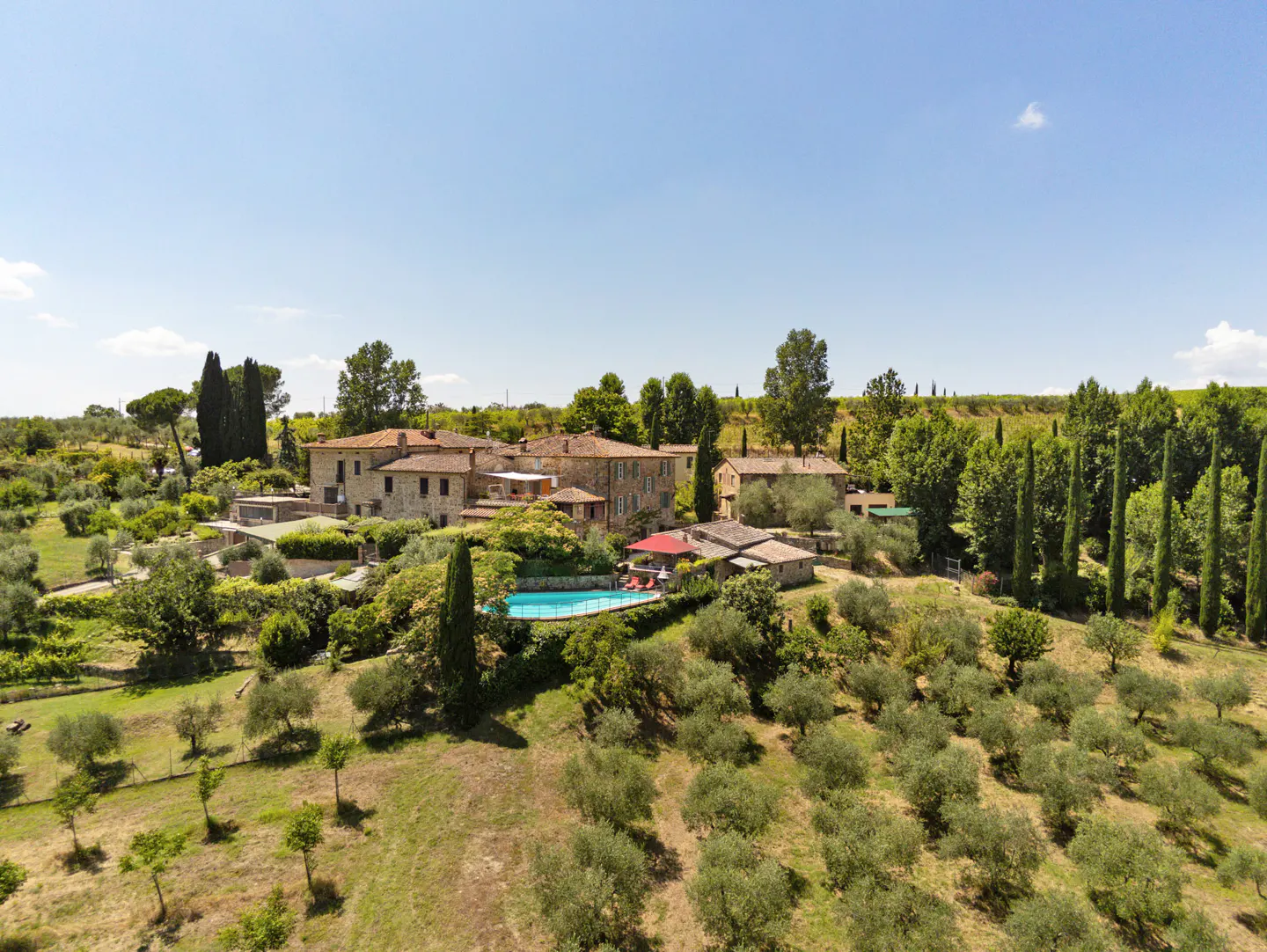 Aerial view of a Tuscan villa with a pool, surrounded by olive trees and cypress trees under a blue sky.