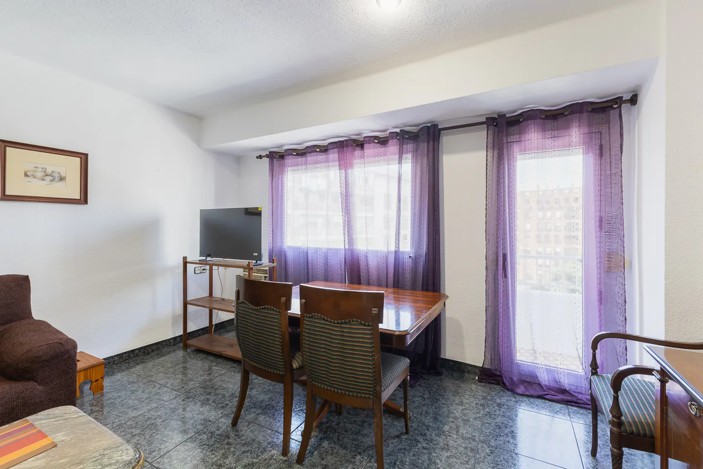 Dining area with a wooden table, two striped chairs, a TV on a shelf, and purple curtains leading to a balcony.