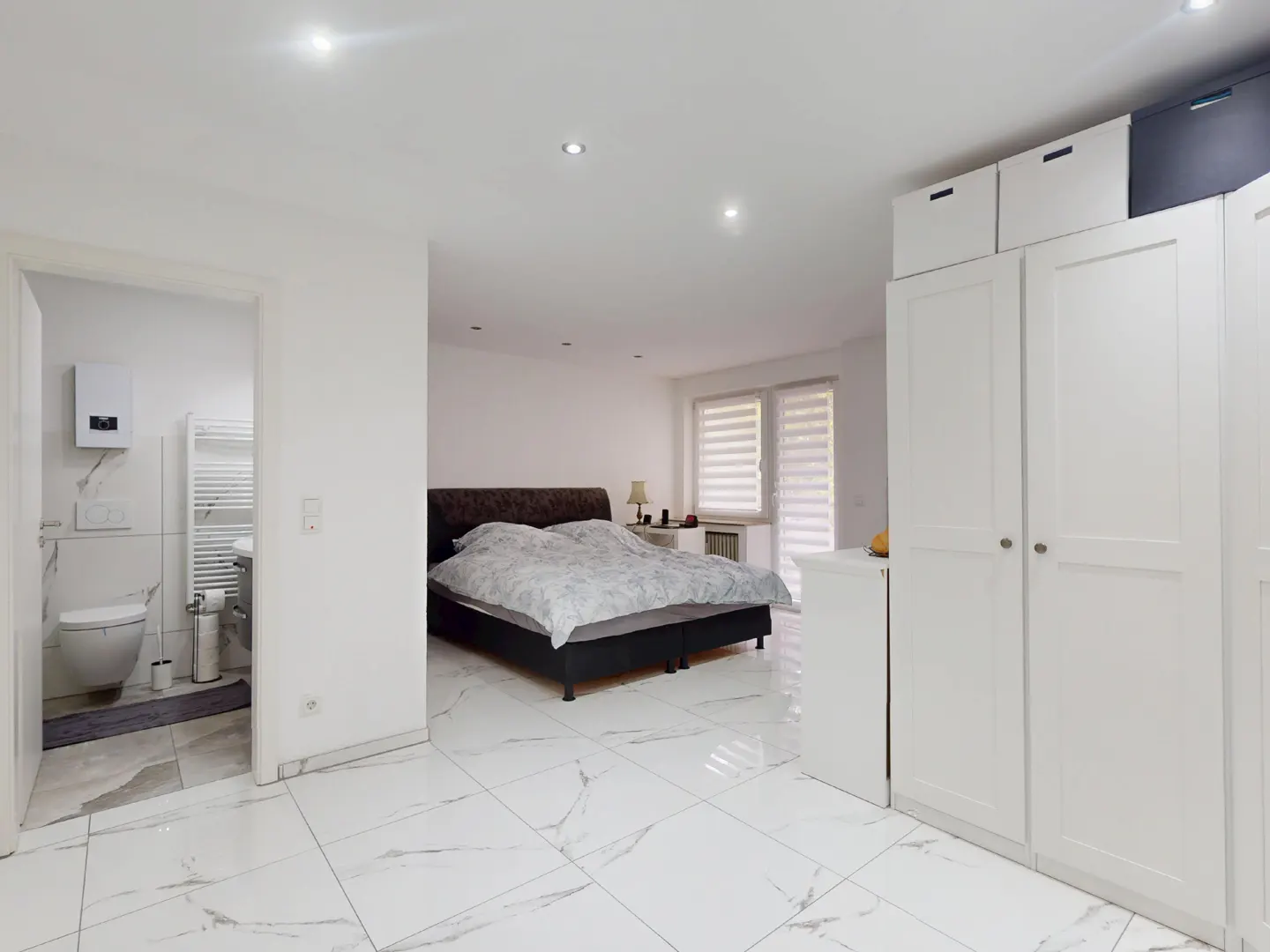Bright bedroom with white marble floors, a bed, and a bathroom visible through a doorway. White wardrobes stand to the right.