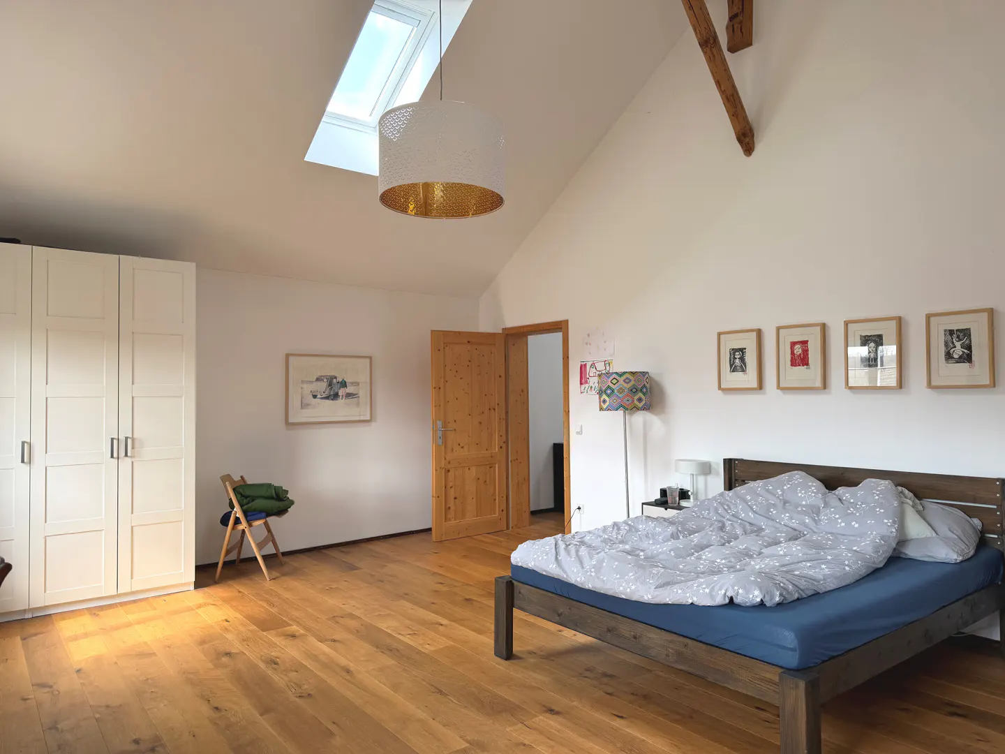 Bedroom with wood floors, white walls, and a skylight. A bed with blue sheets and a patterned duvet is on the right. There is a white wardrobe on the left.