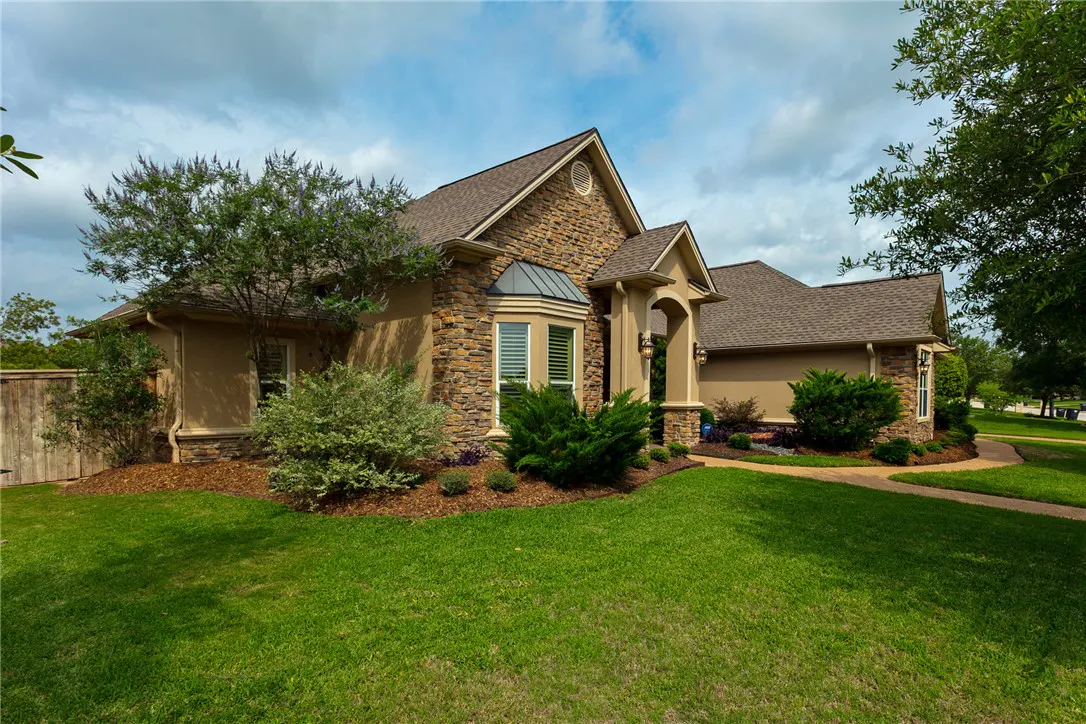 A tan and stone house with a brown roof sits on a green lawn under a cloudy sky. Bushes and trees surround the house.
