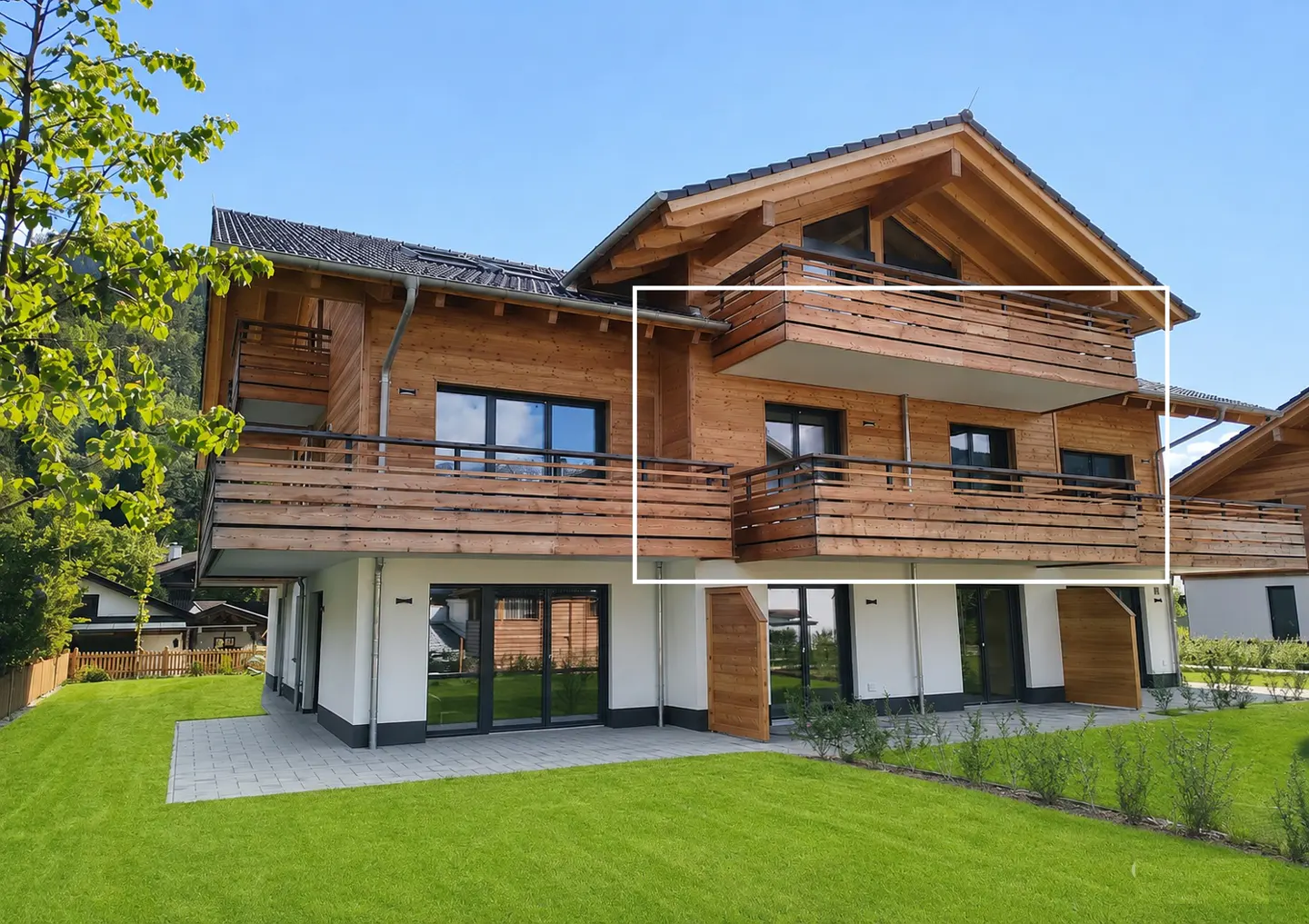 Exterior view of a modern, two-story wood house with a balcony, white base, and green lawn under a clear blue sky.