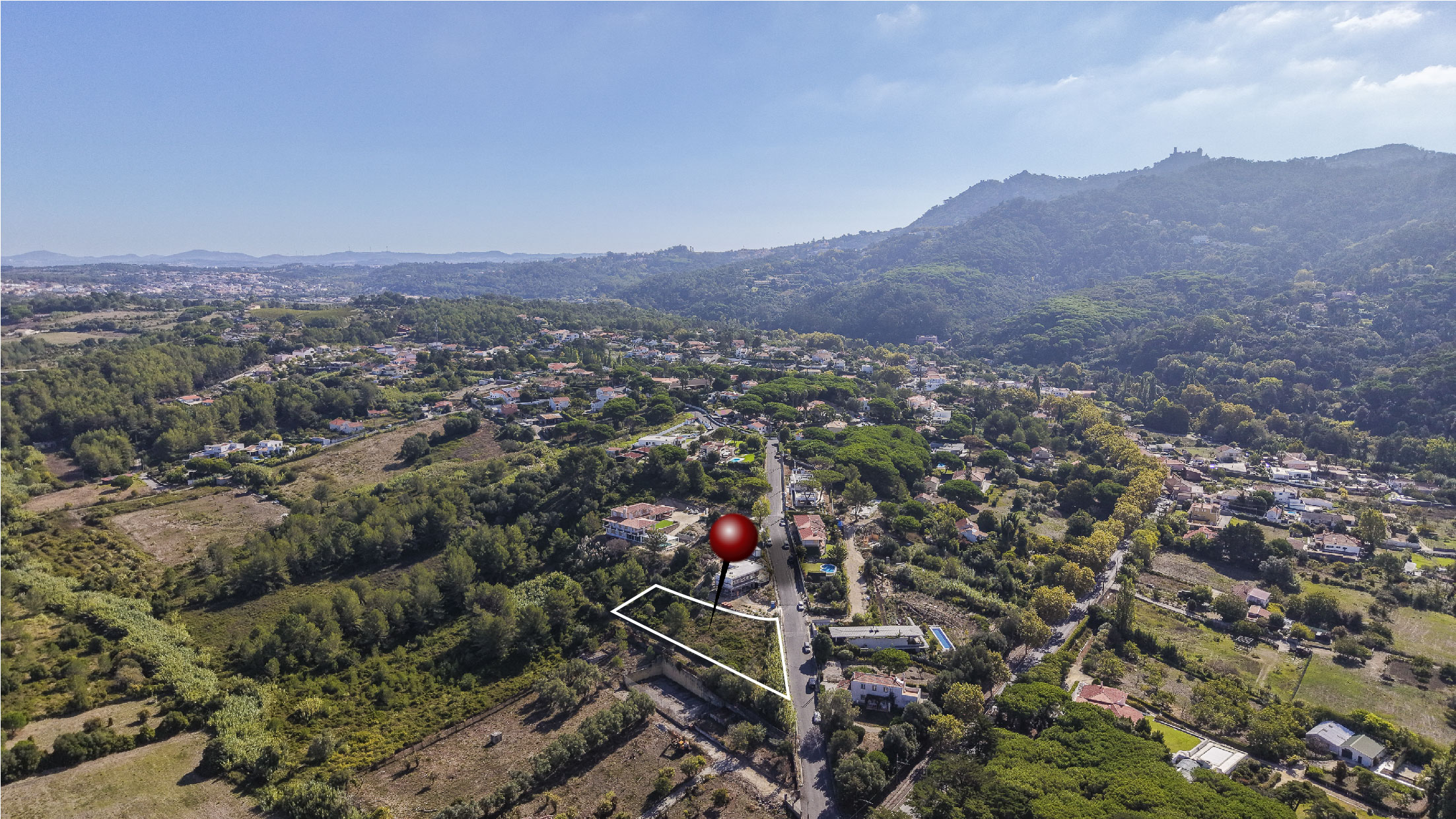 Aerial view of a hillside plot outlined in white with a red location pin, set in a lush, mountainous residential area. [AI-generated text]