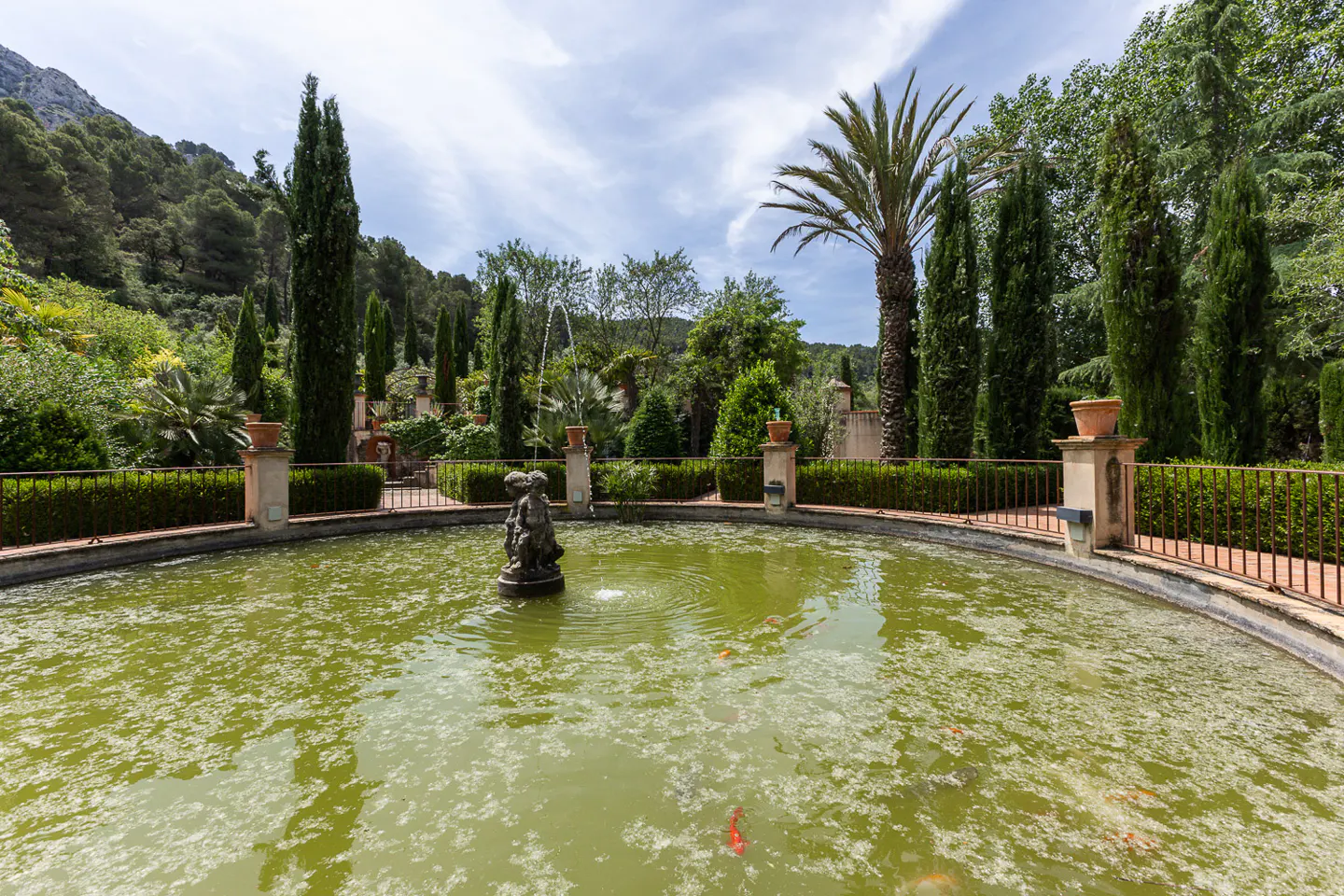 A circular pond with a central fountain statue, surrounded by lush green trees, hedges, and a metal fence under a blue sky.