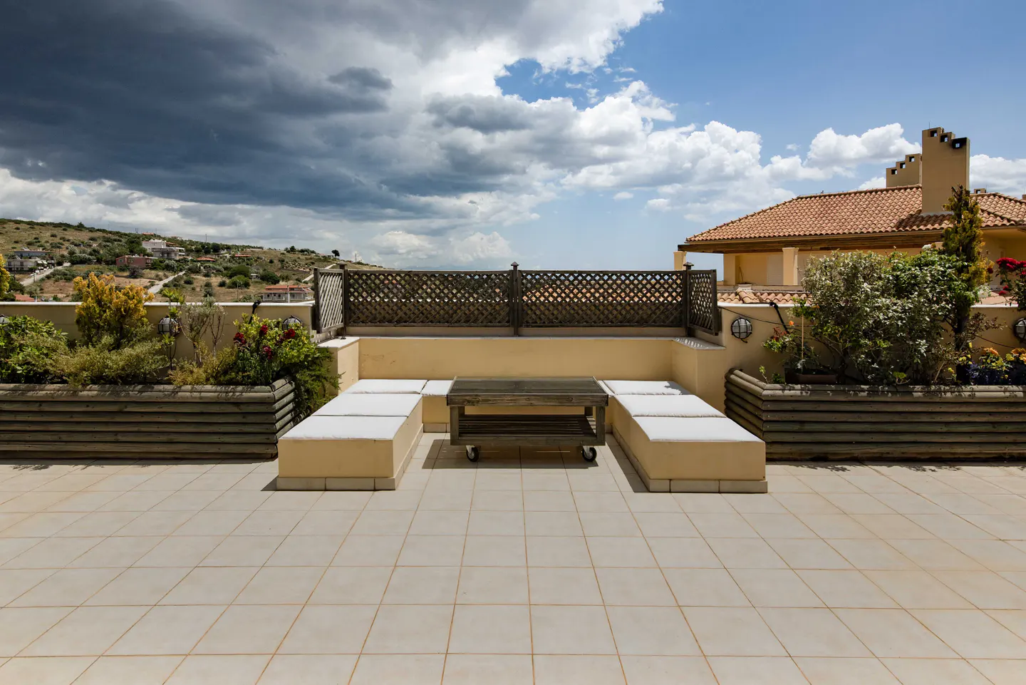 A rooftop patio with a tiled floor, built-in seating, a wooden table, and a lattice railing overlooking a landscape.