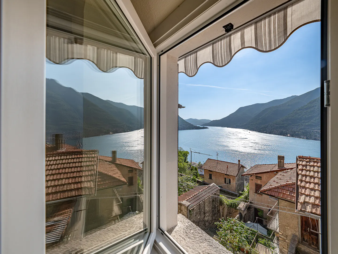 Open white window frames a scenic view of a lake, mountains, and terracotta-roofed buildings under a blue sky.