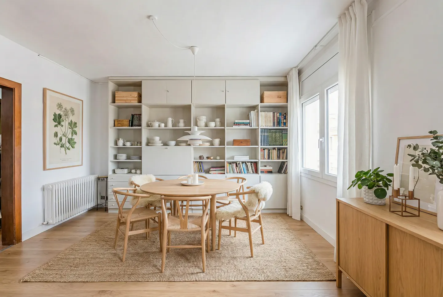 Bright dining room with a round wooden table, six chairs with sheepskin throws, and a large built-in bookshelf filled with books and dishes.
