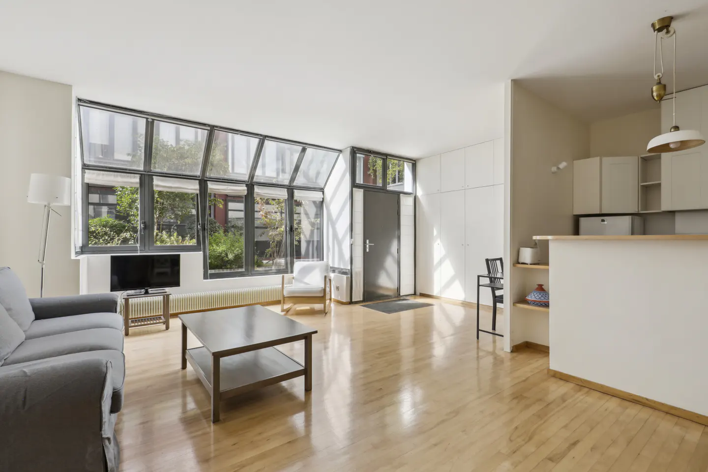 Bright living room with hardwood floors, a gray sofa, coffee table, and large windows overlooking a courtyard. A kitchen area is visible in the background.