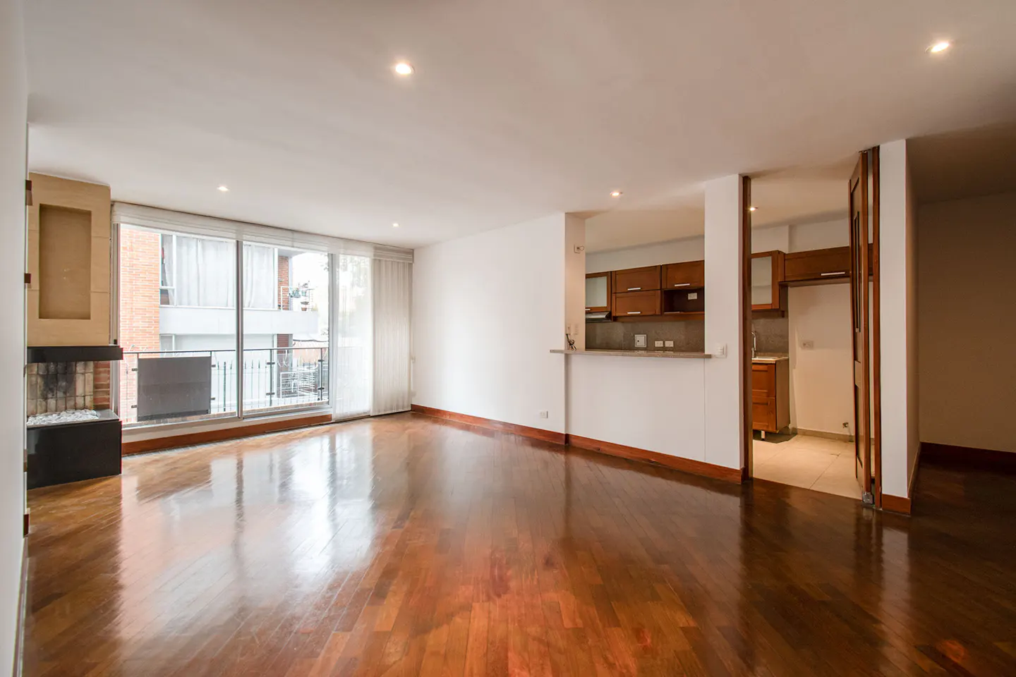 Bright, empty living room with hardwood floors, a fireplace, and a balcony. The kitchen is visible through an opening.