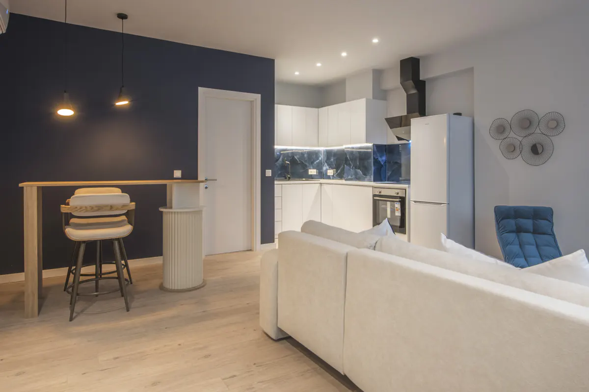 A modern apartment interior with a white sofa, kitchen, and bar area. The walls are navy and light gray.