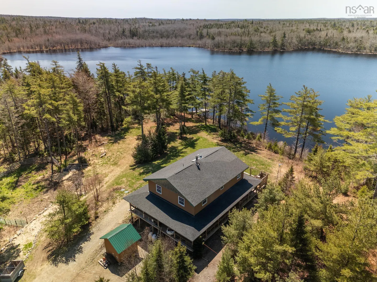 Aerial view of a brown two-story house with a dark roof, surrounded by green trees and a blue lake in the background.