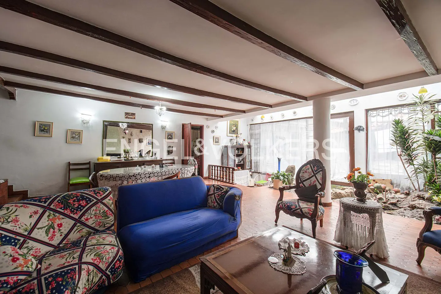 A living room with a blue sofa, floral armchairs, and a wooden coffee table. The ceiling has exposed wooden beams. A garden is visible through the window.