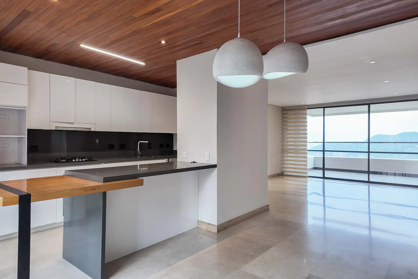 A modern kitchen with white cabinets, a dark countertop, and a wooden ceiling. Two white pendant lights hang over the island. Large windows offer a view of mountains.