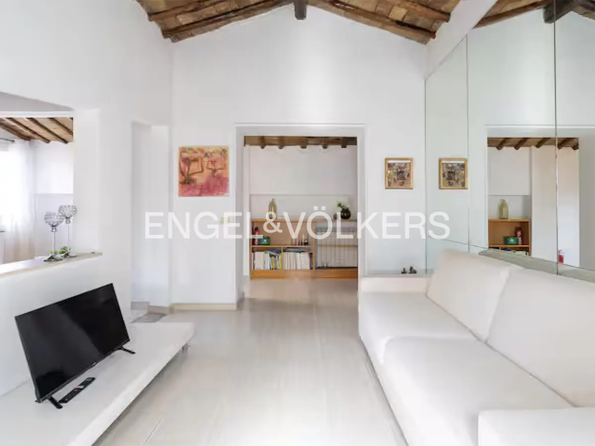 Bright living room with white walls, tile floor, and exposed wood beam ceiling. A white sofa faces a TV. Artwork adorns the walls.