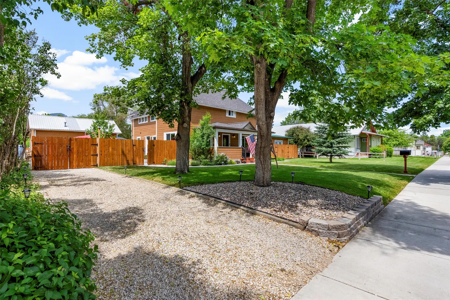 A two-story orange house with a gravel driveway, green lawn, and trees on a sunny day. An American flag is on the porch.