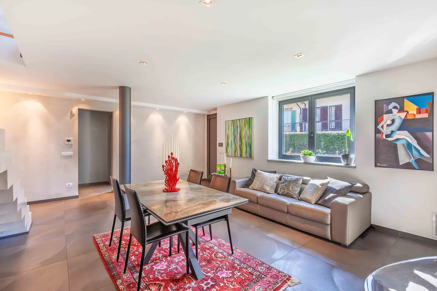 Bright living room with a dining table, gray sofa, and modern art. A red rug adds warmth to the gray tile floor. Natural light streams through a window.