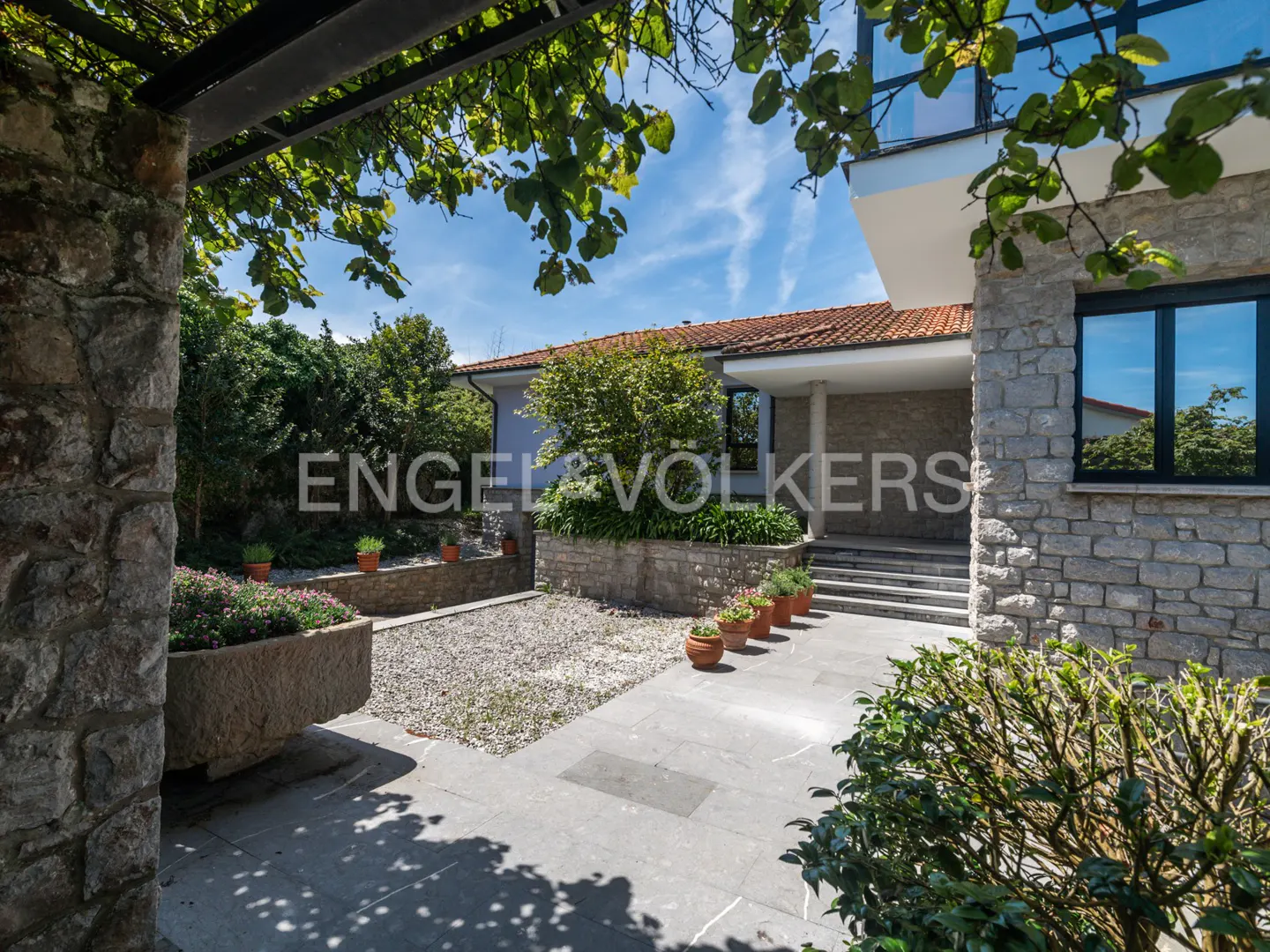 Stone house exterior with a tiled roof, gray stone walls, and a paved patio. Green foliage and potted plants add color. Blue sky visible.