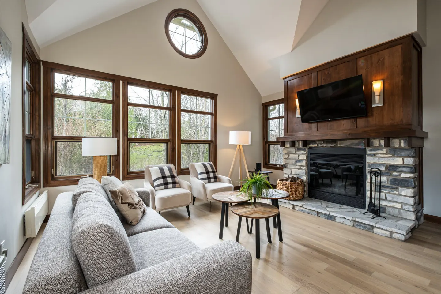 Living room with vaulted ceiling, large windows, stone fireplace, gray sofa, and two armchairs.