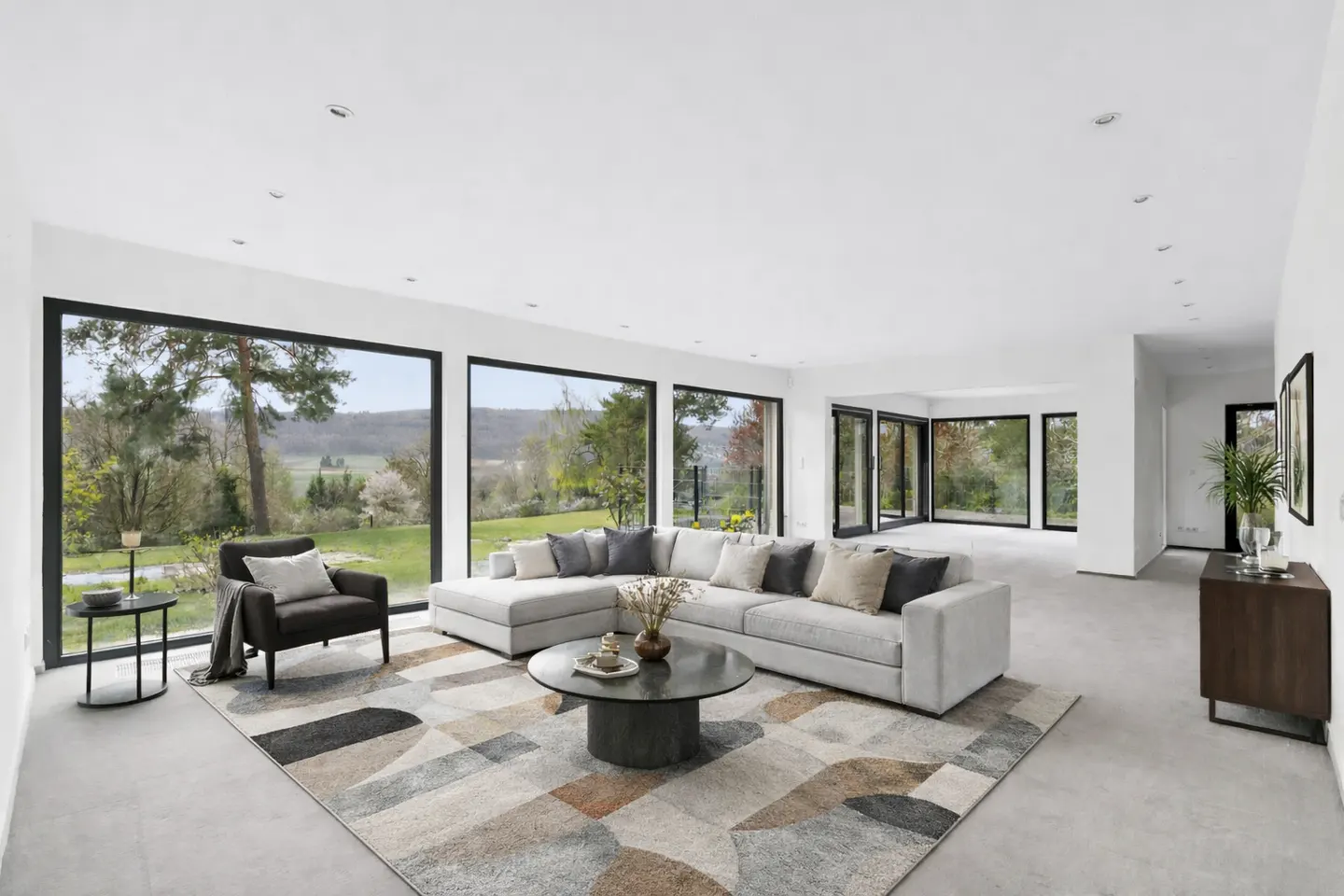 Bright living room with large windows overlooking a green landscape. A gray sectional sofa sits on a patterned rug, with a round coffee table in the center.