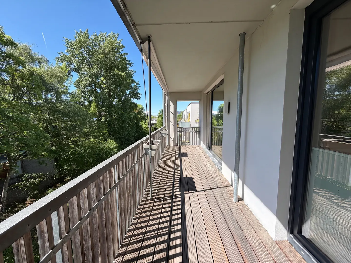 A long, narrow balcony with a wooden floor and railing overlooks lush green trees on a sunny day.