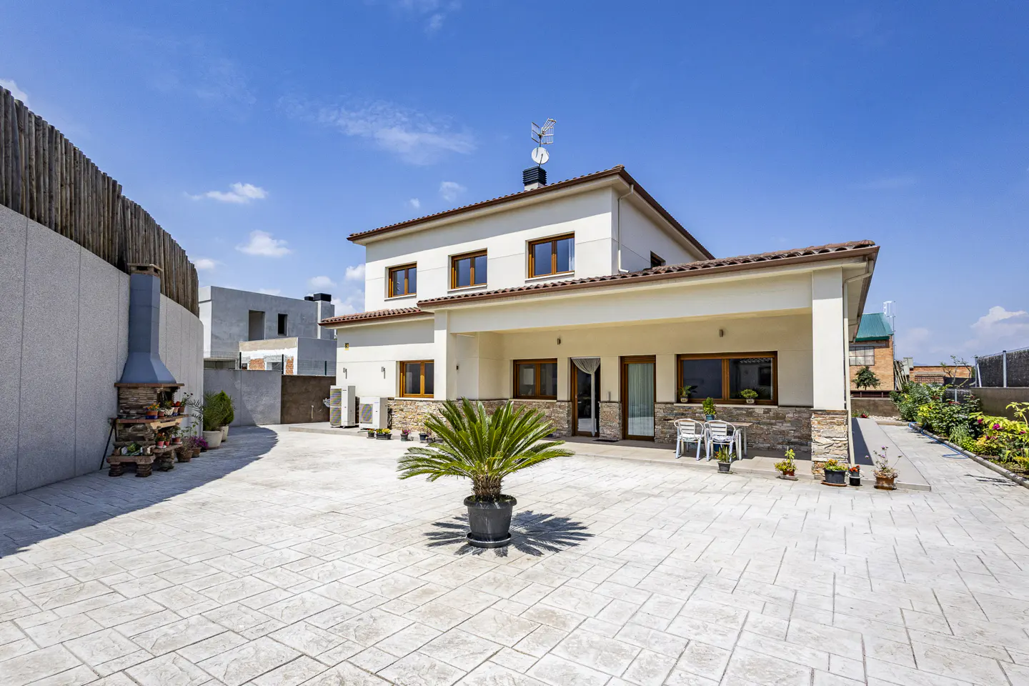 Two-story house with a concrete patio, a grill, and a palm tree in a pot under a blue sky.