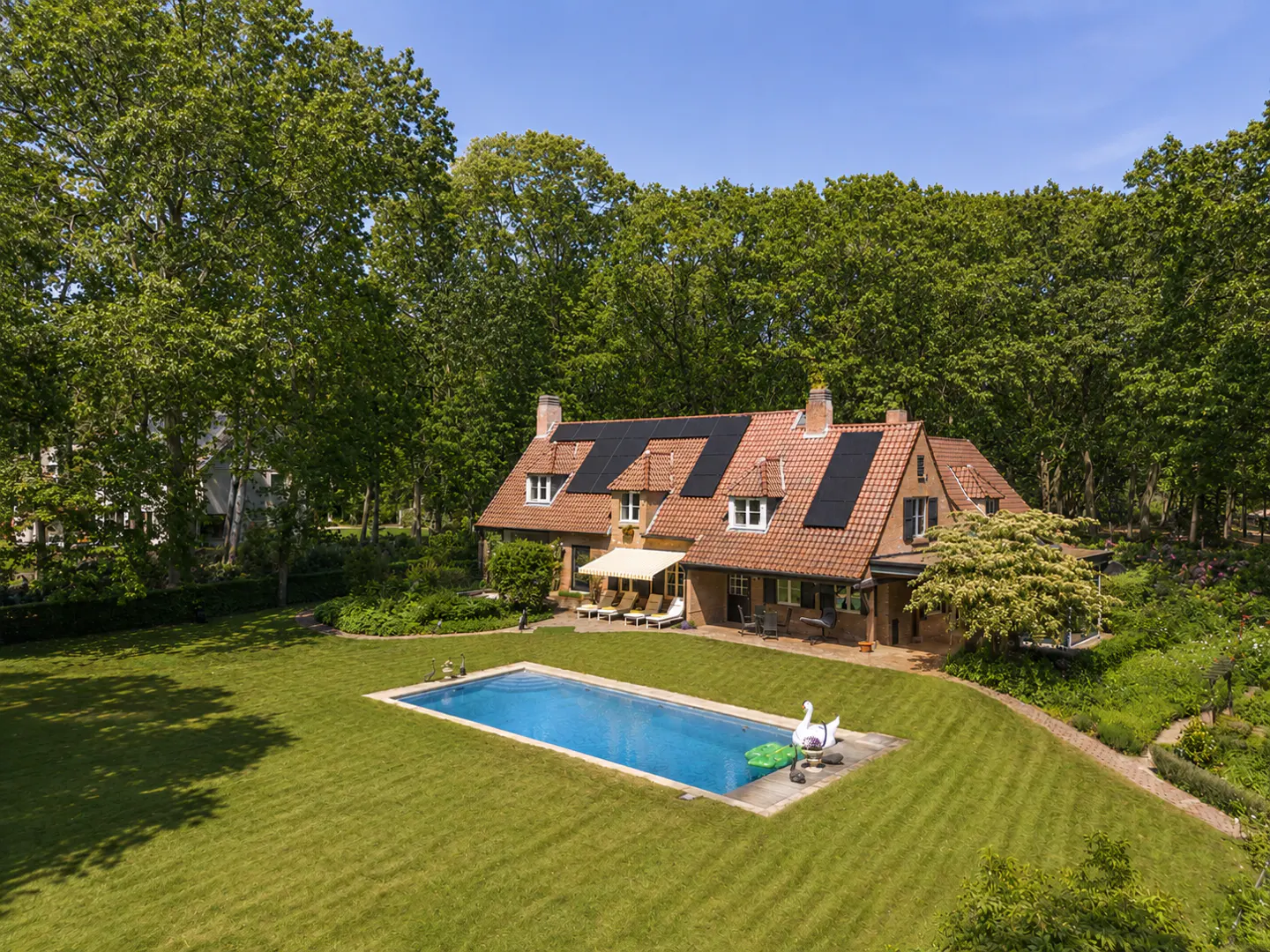 Aerial view of a brick house with a red tile roof, solar panels, a pool, and a large green lawn.