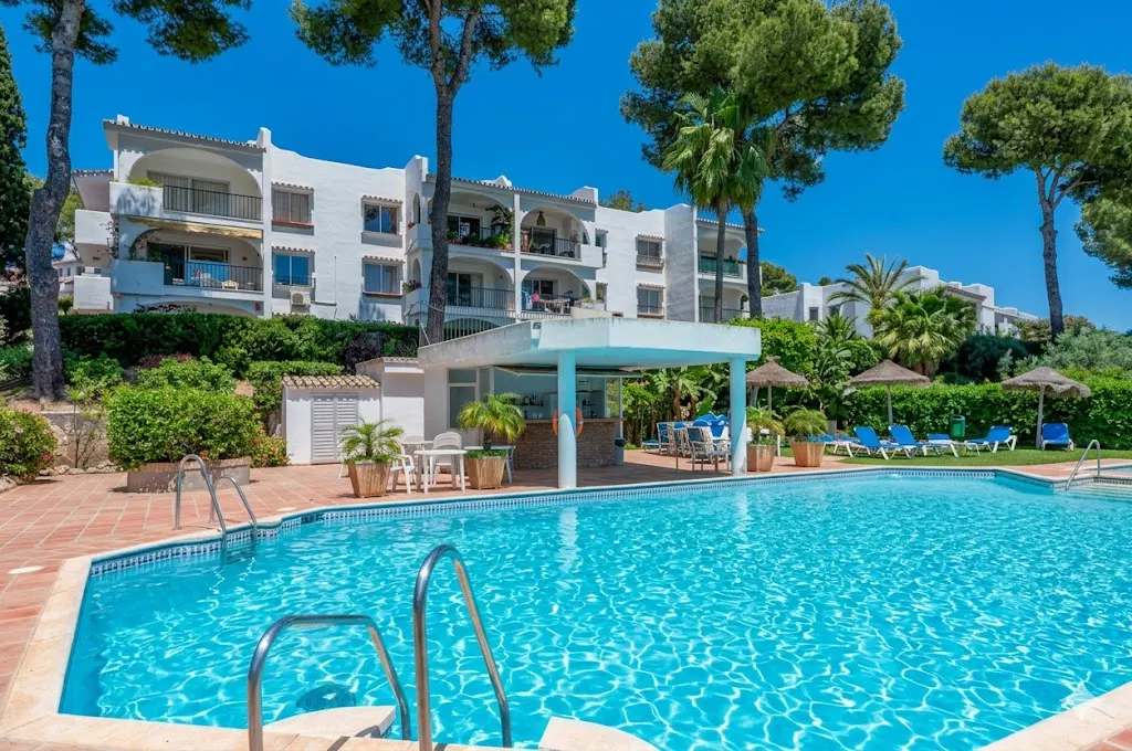 Outdoor pool with turquoise water, bar, and white building with balconies under a clear blue sky. Palm trees and lounge chairs surround the pool.