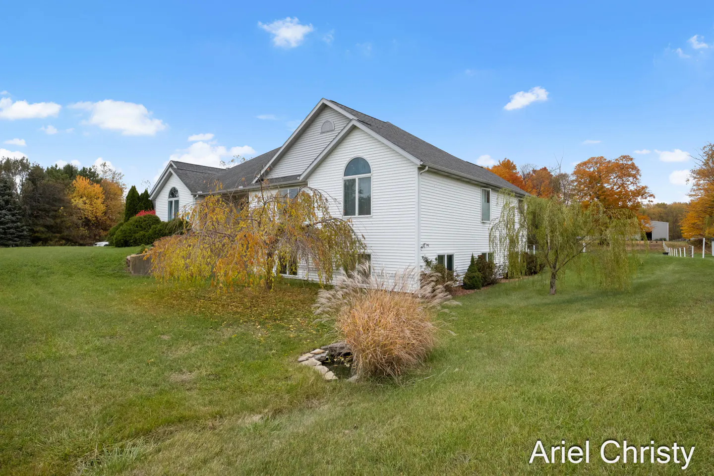 Two-story white house with a gray roof, surrounded by green grass, trees, and a blue sky with white clouds.
