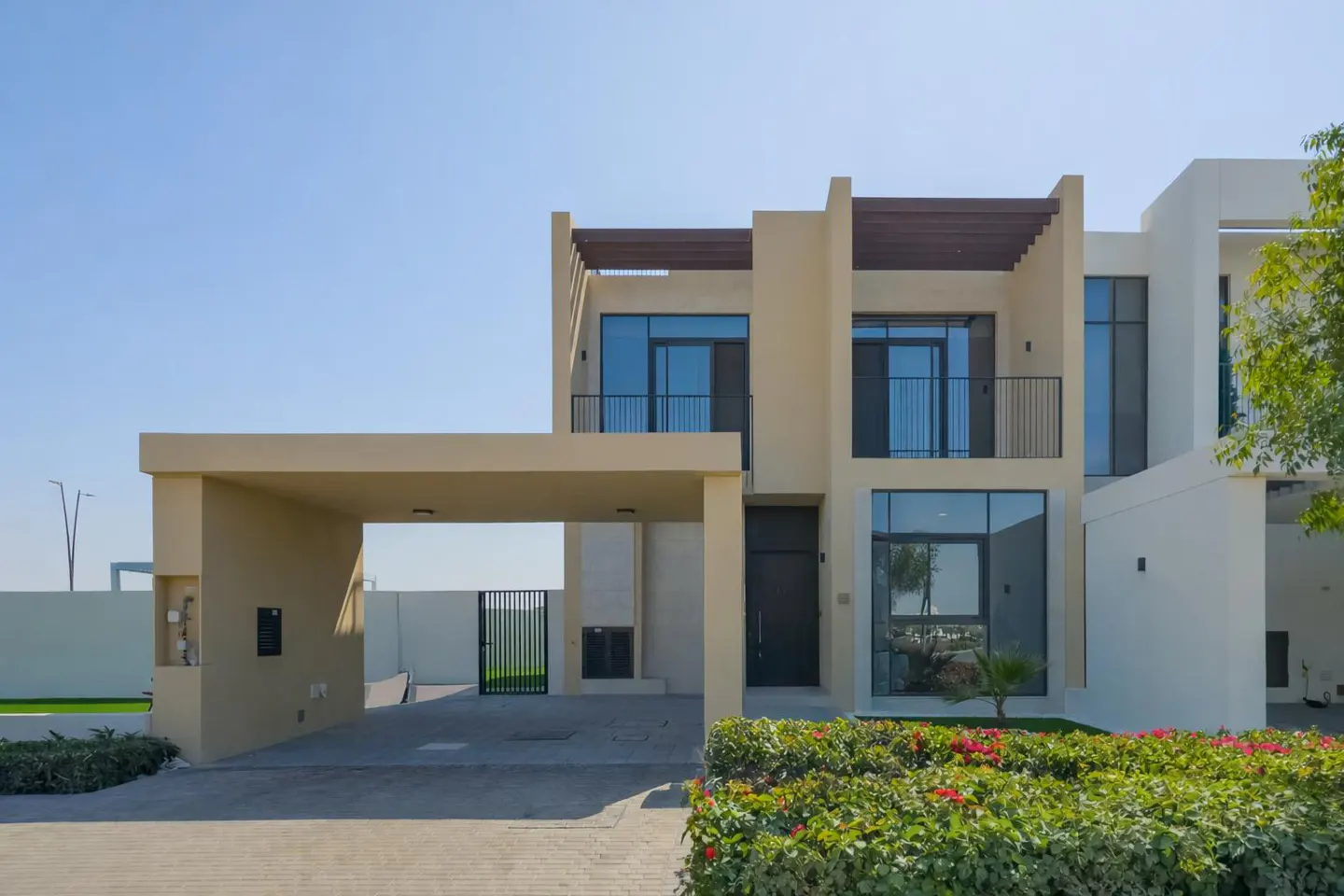 Modern two-story beige villa with a carport, large glass windows, a balcony, and a landscaped garden under a clear sky.