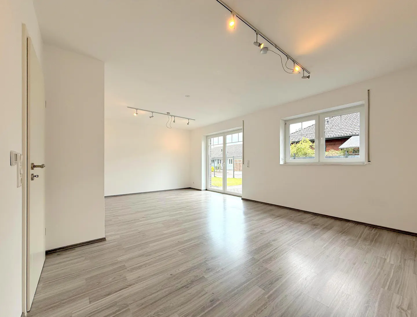 Bright, empty room with gray wood floors, white walls, track lighting, and a window overlooking a yard.