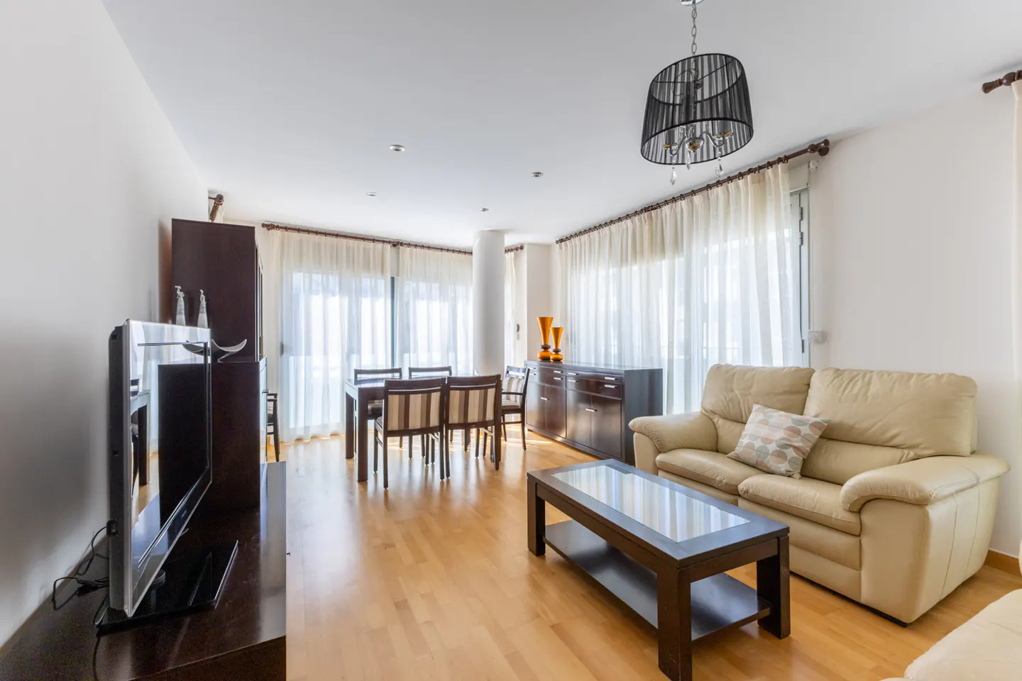 Bright living room with wood floors, a beige sofa, a dark wood coffee table, and a dining table with chairs.