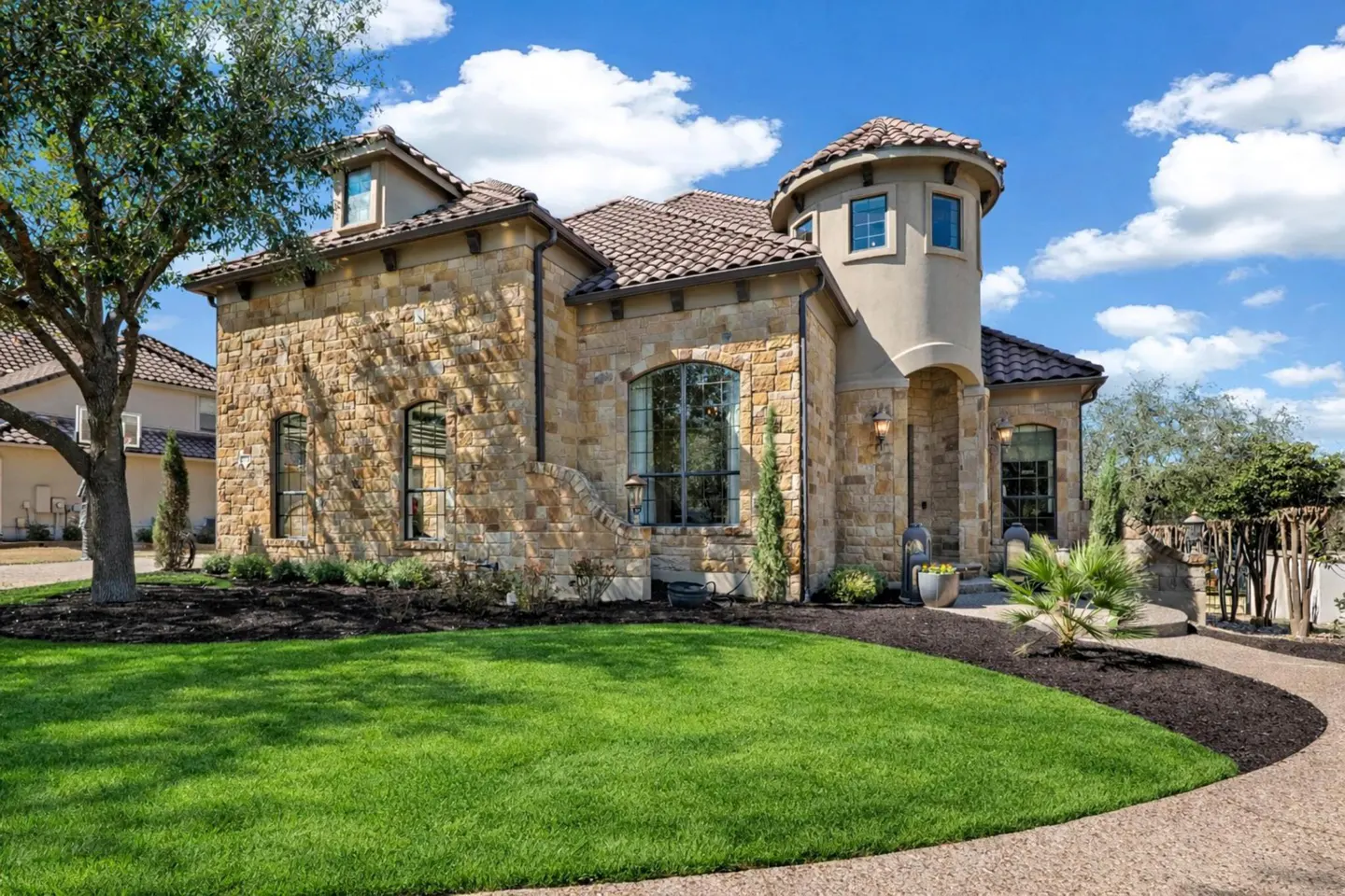 A tan stone house with a turret, a brown tile roof, and a green lawn under a blue, cloudy sky.