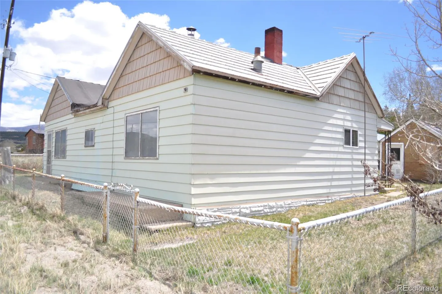 Exterior view of a light green, single-story house with a red brick chimney and a chain-link fence in a grassy yard.