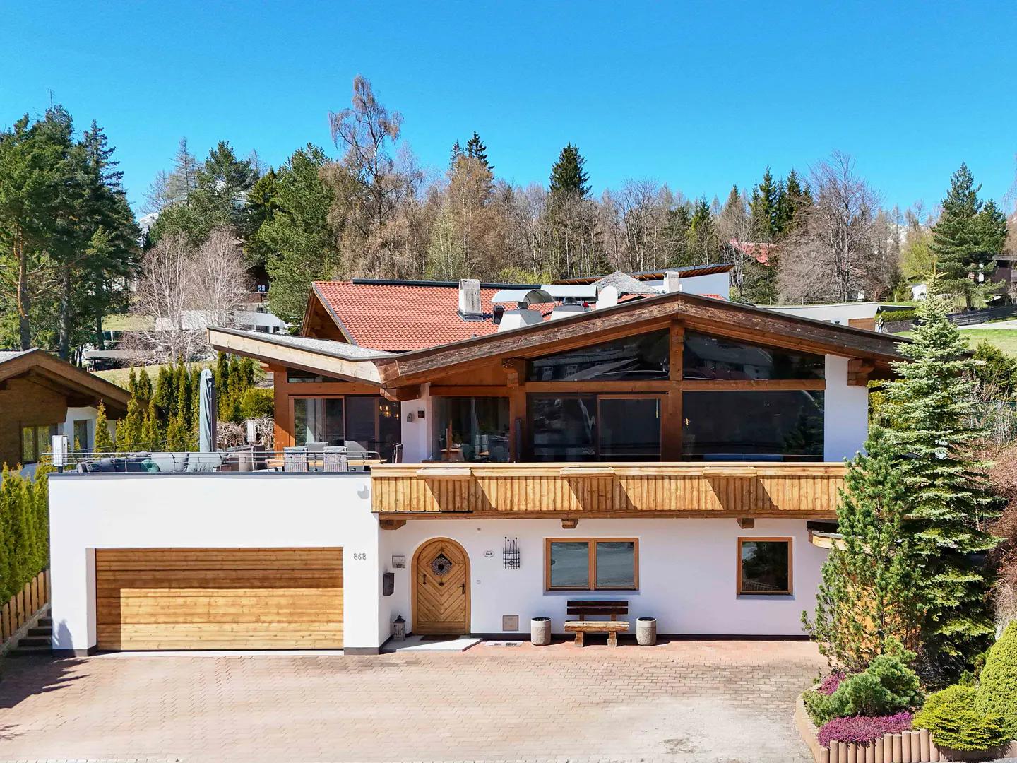 Two-story white house with wood trim, a red tile roof, and a wood garage door, set against a backdrop of green trees and a blue sky.