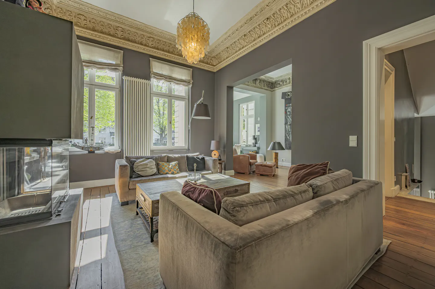 Living room with gray walls, hardwood floors, and ornate ceiling trim. Two sofas, a chandelier, and large windows provide natural light.