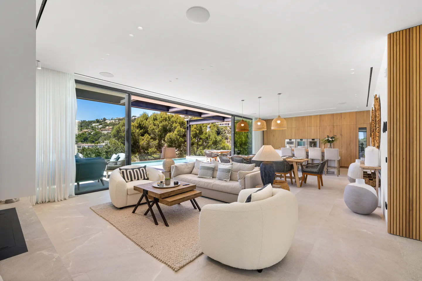 Bright, open-plan living space with white sofas, wood accents, and a view of a pool and lush greenery through large sliding glass doors.