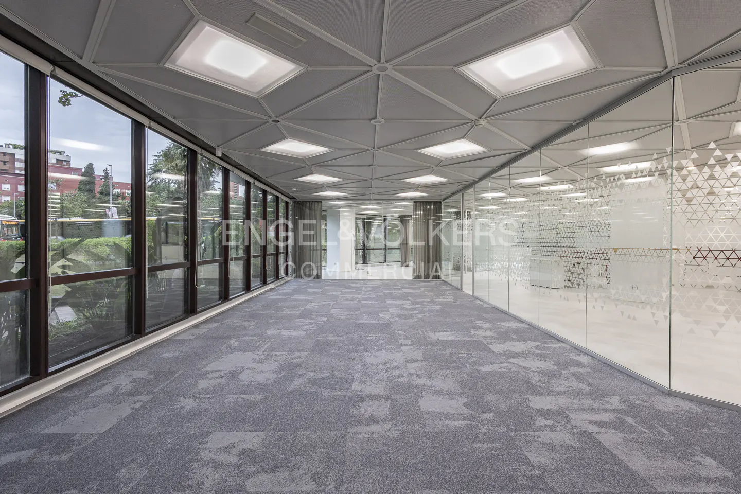 Empty office space with gray carpet, glass walls with geometric patterns, and large windows with a view of trees and buildings.