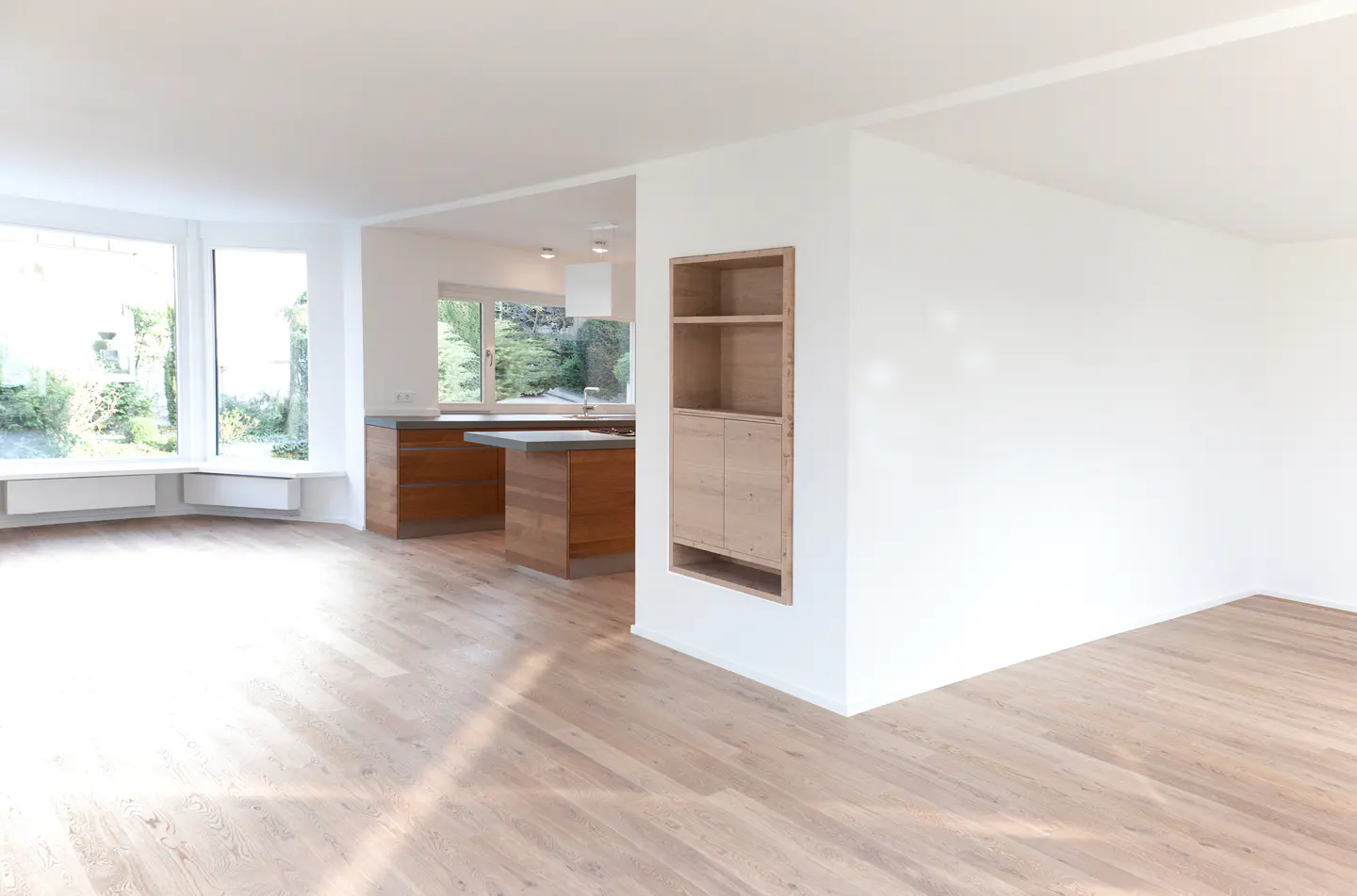 Bright, open-concept living space with light wood floors, white walls, and large windows. A modern kitchen is visible in the background.