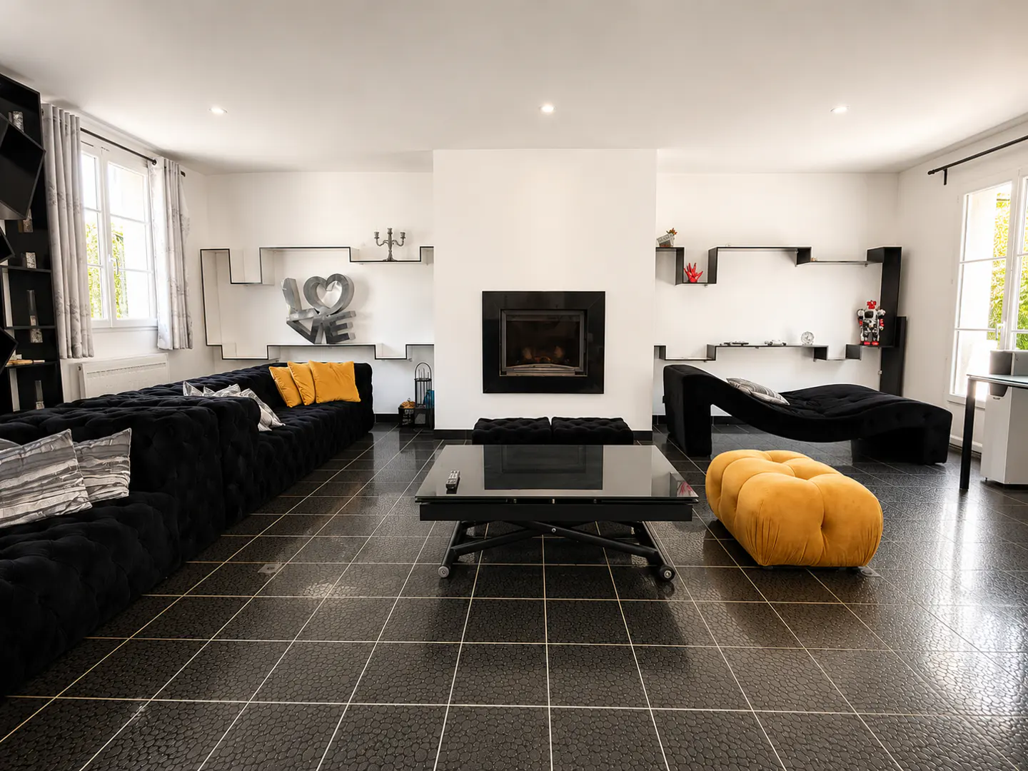Modern living room with black tufted sofa, yellow ottoman, fireplace, and black tiled floors in a bright, white space.