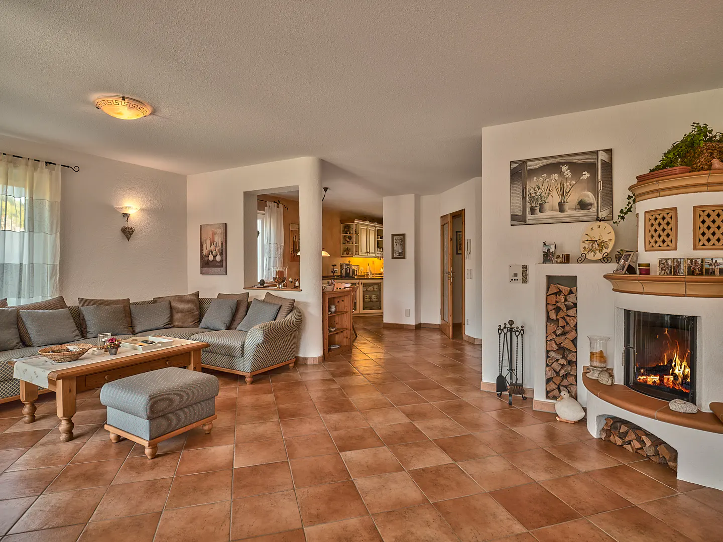 Living room with brown tile floor, a gray sectional sofa, and a white fireplace with a fire burning. A kitchen is visible in the background.