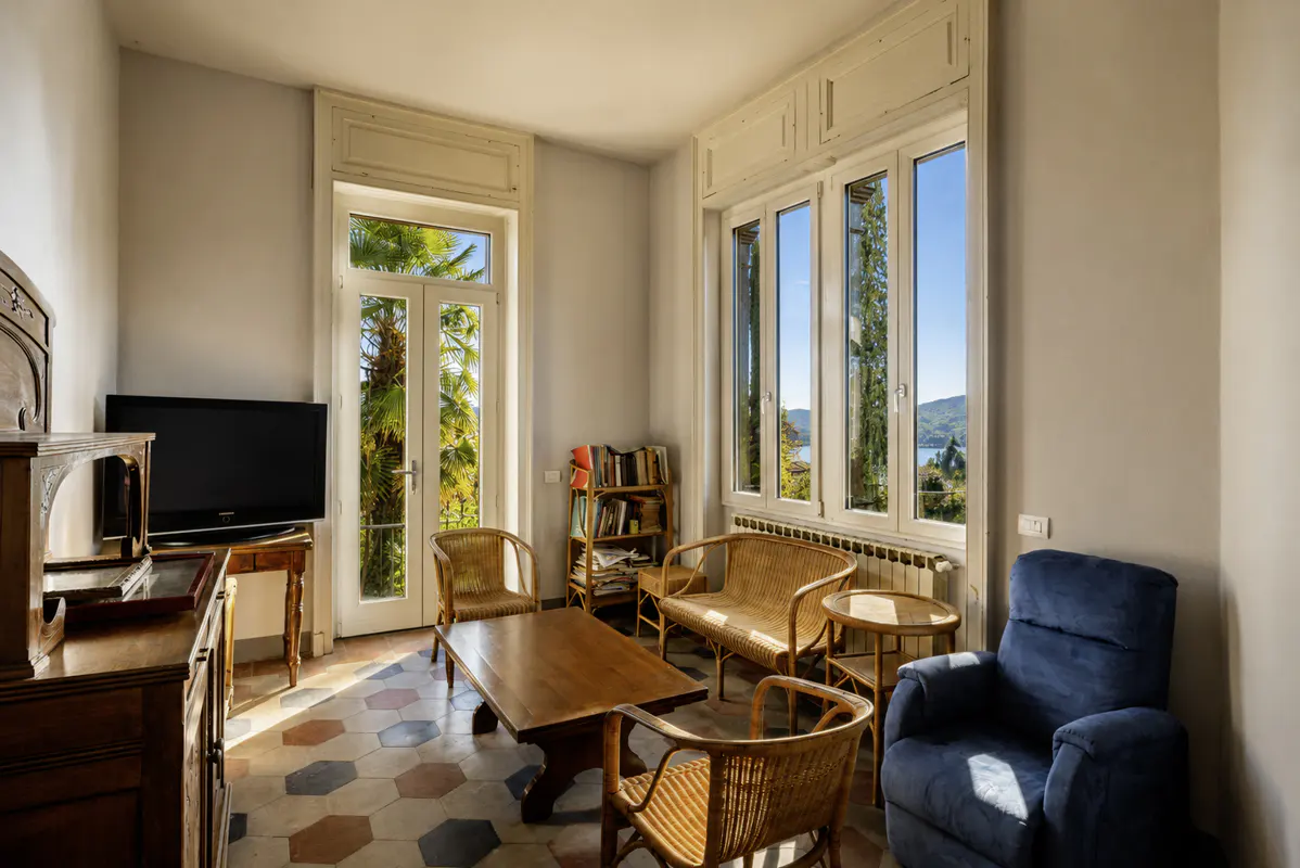 Bright living room with wicker furniture, a blue armchair, and a wooden coffee table on a patterned tile floor. Large windows offer a view of trees and water.