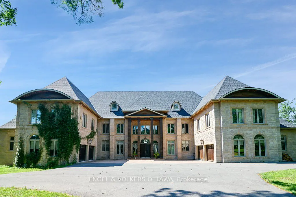 Beige stone mansion with a gray roof, symmetrical design, and a large driveway under a blue sky.