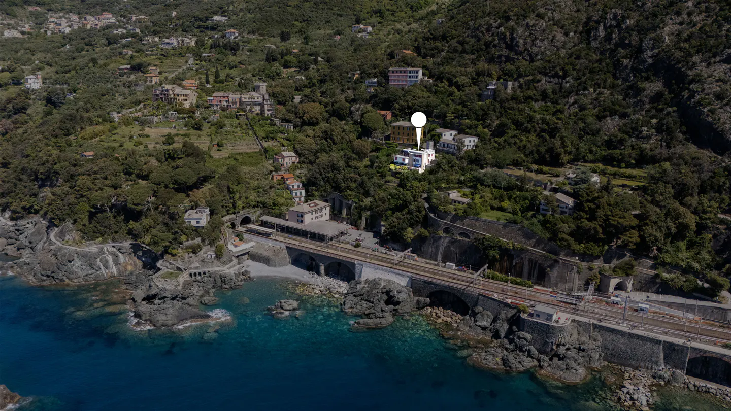 Aerial view of a white house with a location pin, nestled on a green hillside near a train track and the blue sea.