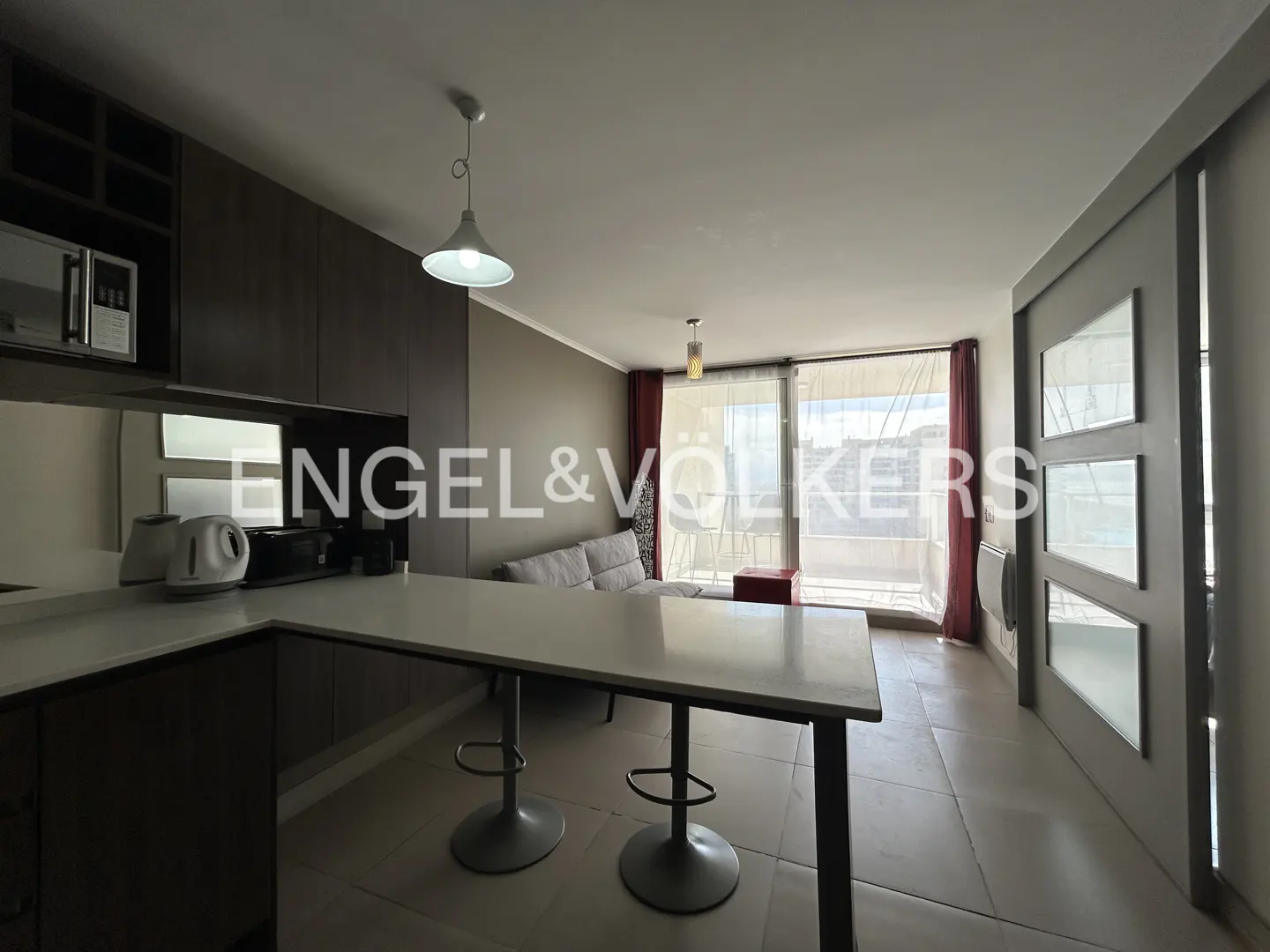 A modern apartment interior with a kitchen island, bar stools, and a view from the balcony.