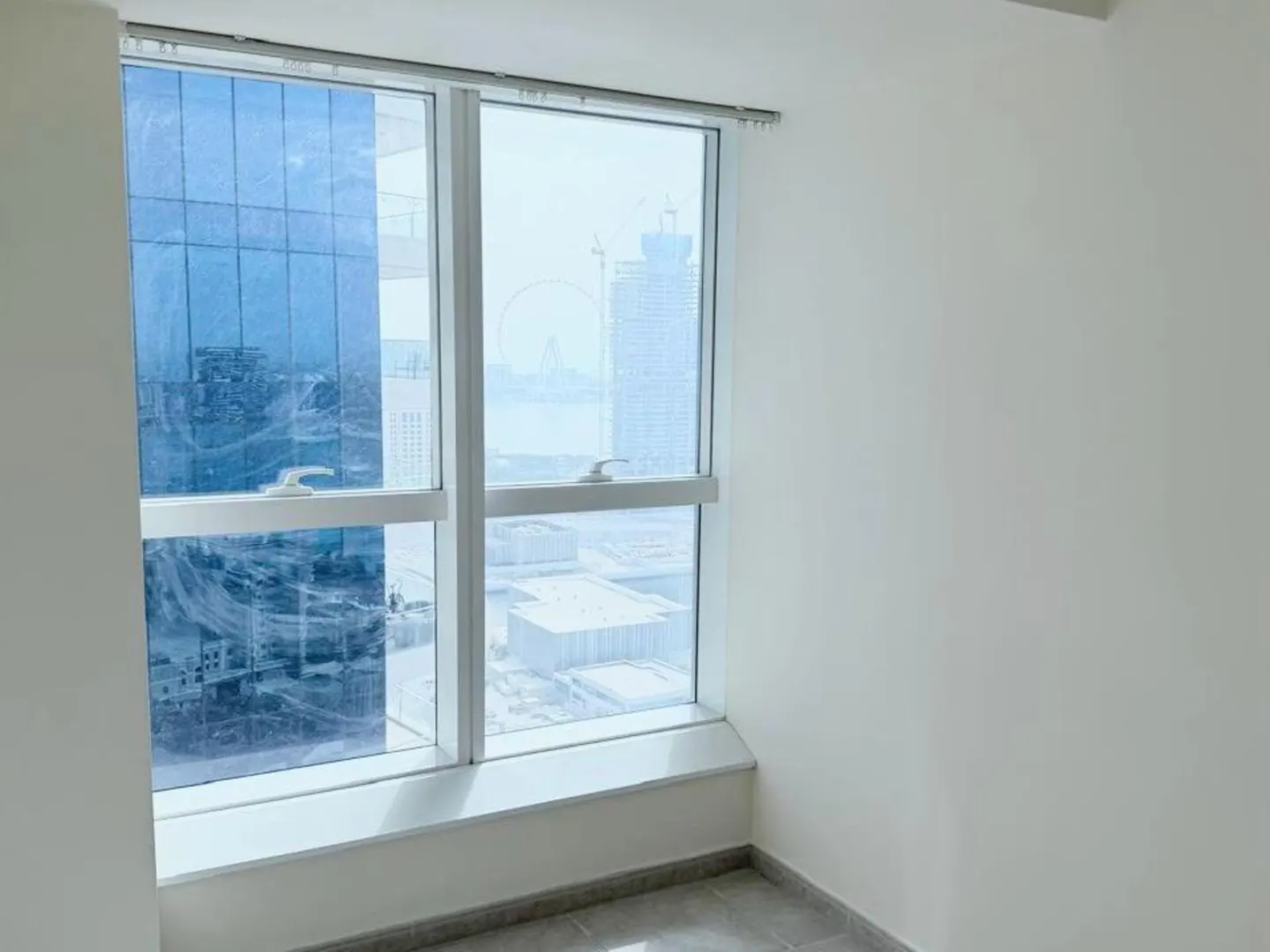 An empty room with white walls and gray tile floors features a large window with a city view.
