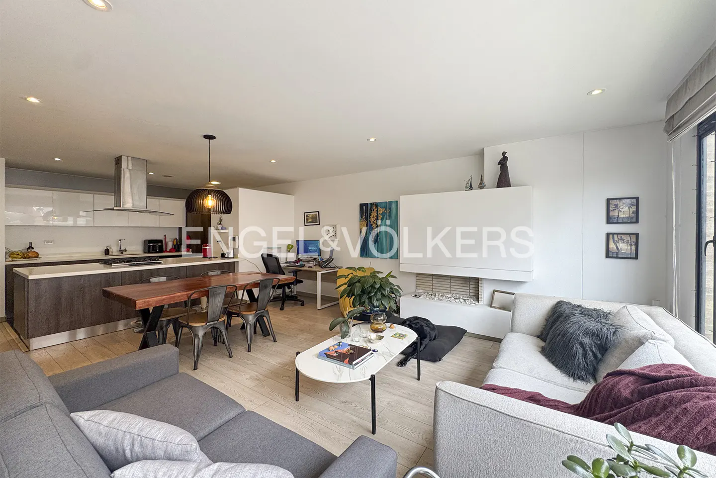 Open-concept living space with a gray sofa, dining table, kitchen island, and a white fireplace. A black dog rests near the sofa.