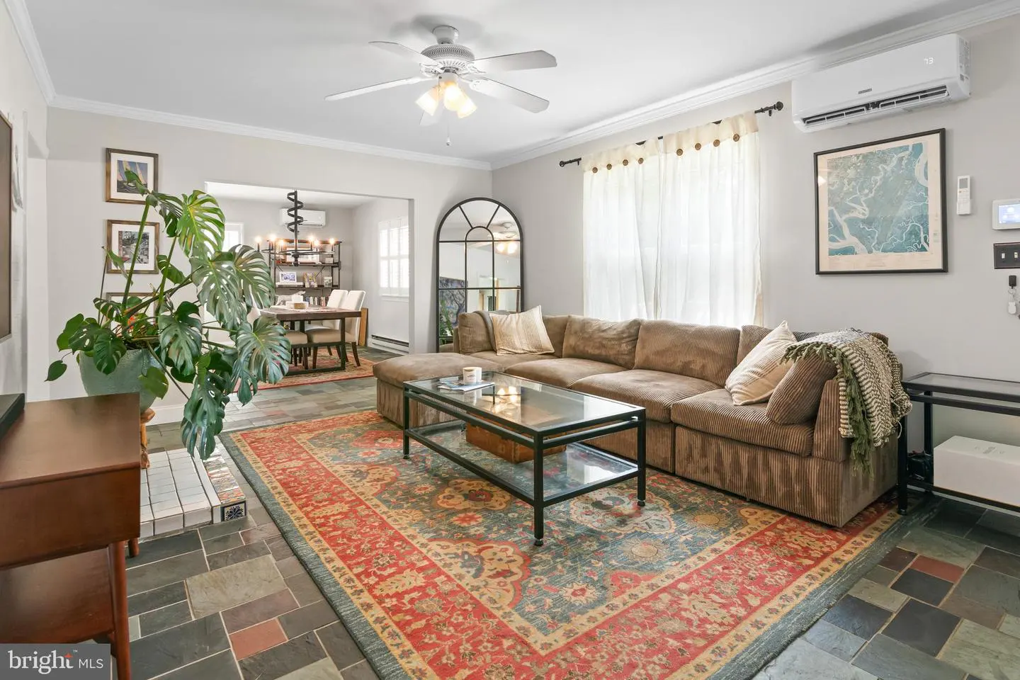 Living room with brown sectional sofa, glass coffee table, large patterned rug, and a tall plant on a slate tile floor.
