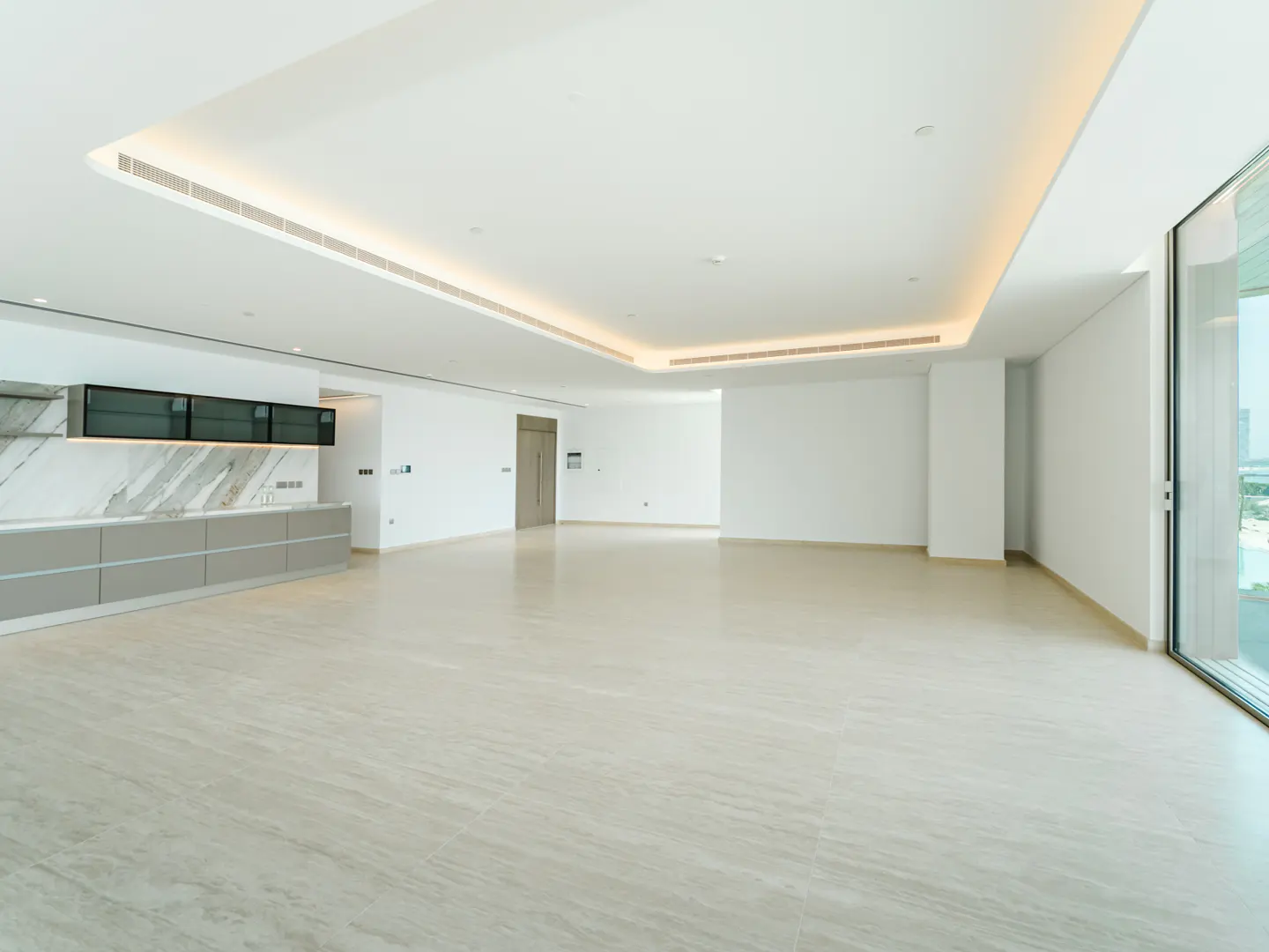 Bright, empty room with marble floors, white walls, and recessed lighting. Kitchenette with gray cabinets and marble backsplash on the left. Large window on the right.
