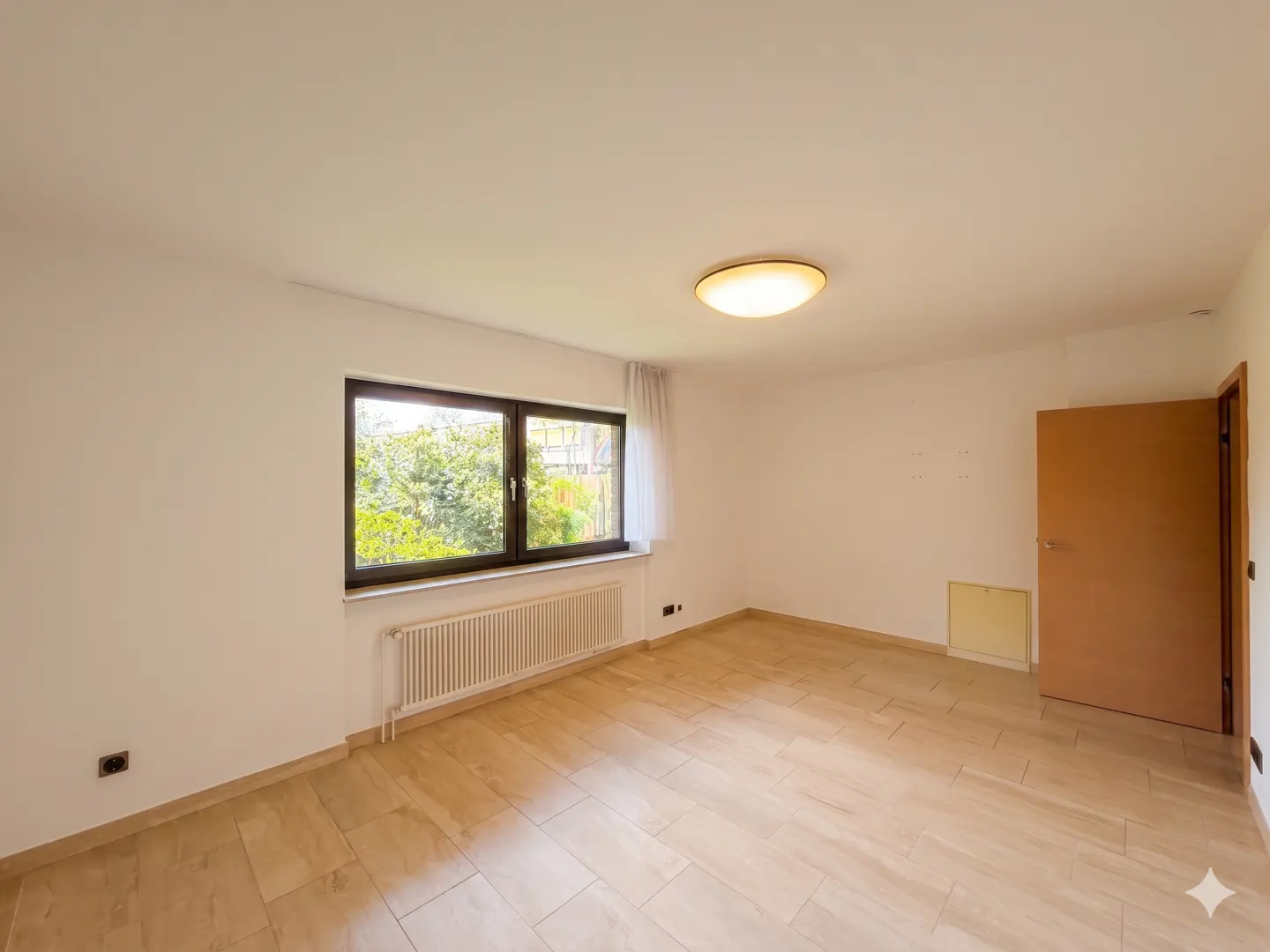 Bright, empty room with tan tile floor, white walls, and a brown door. A window shows green trees outside. A radiator sits under the window.